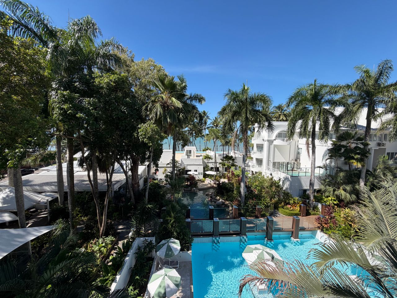 Aerial view of a tropical resort featuring palm trees, swimming pool, and ocean view under clear blue sky.